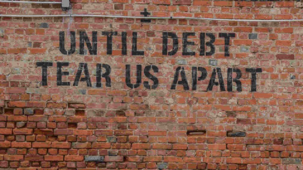 Brick wall spray painted with "Until Debt Tears Us Apart" on red brick wall.